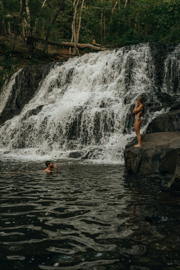 Destination Wedding and Elopement Photographer | Aisha Lee Photography Cinematic Wedding Photographer | Two people by a waterfall in a forested area. One person stands on a rock, the other swims, both in swimwear.