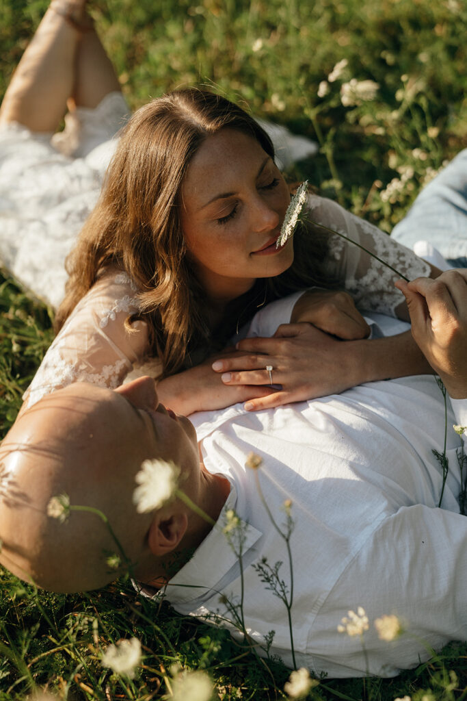 Moody Wedding Photographer Rhode Island | Aisha Lee Photography Cinematic Wedding Photographer | A couple lies in a grassy field, embraced closely. The woman, in a lace dress, gently sniffs a flower