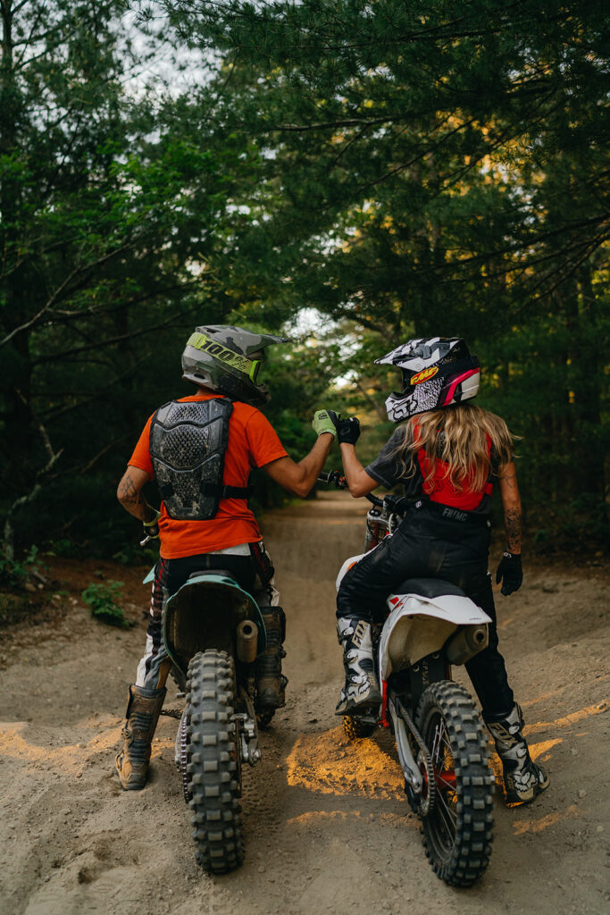 Destination Engagement Photographer New England | Aisha Lee Photography Cinematic Wedding Photographer | A couple motorcyclists in protective gear fist bump on dirt bikes in a forest trail.