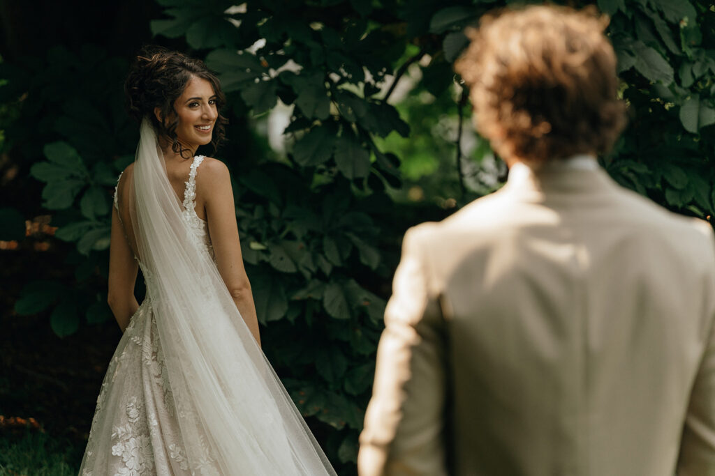 Bride smiles toward her partner beneath deep green foliage during an intimate first-look moment, highlighting the beauty of wooded wedding venues in New Hampshire.