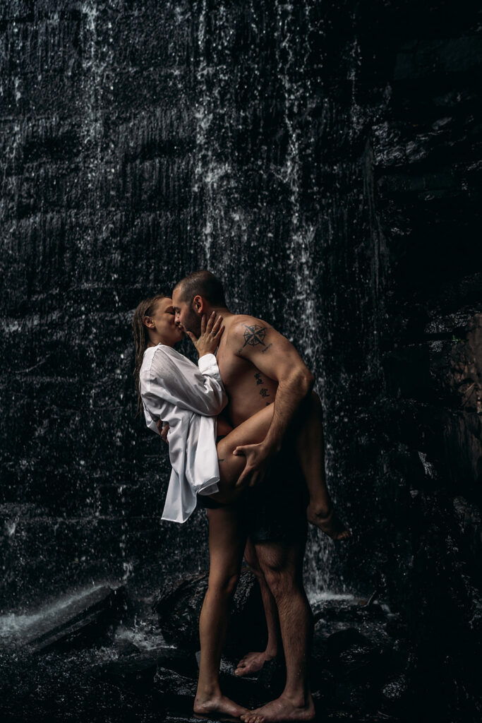 Destination Engagement Photographer New England | Aisha Lee Photography Cinematic Wedding Photographer | A couple embraces passionately under a cascading waterfall. The woman wears a white shirt, while the man is shirtless.
