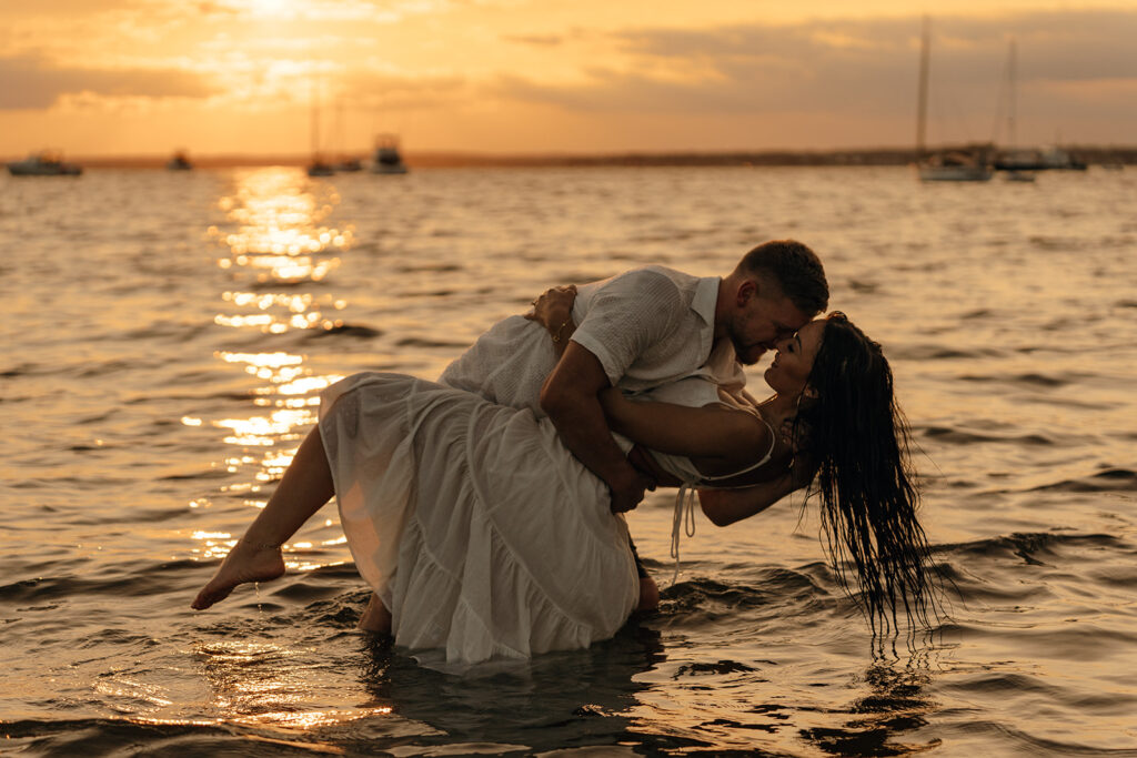 Couple sharing a romantic dip in shallow ocean water at sunset during a Rhode Island beach wedding