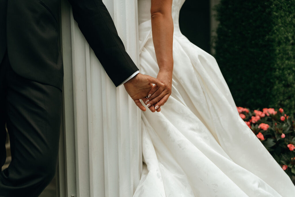 Castle Wedding Venues in New England | Aisha Lee Photography Cinematic Wedding Photographer | Close-up of a bride and groom holding hands around a white column. The bride wears a white wedding dress, and the groom is in a black suit.