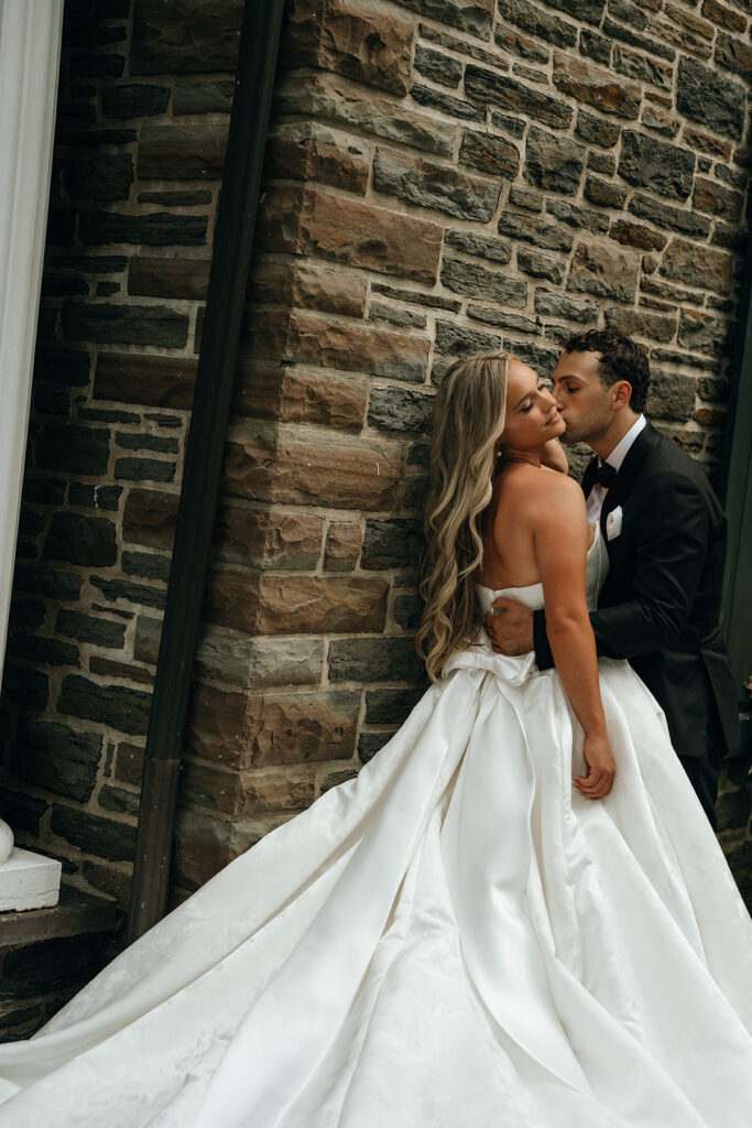 Castles in New England | Aisha Lee Photography Cinematic Wedding Photographer | A groom in a black tuxedo kisses a smiling bride wearing a flowing white gown in front of a rustic stone wall. The mood is romantic and intimate.