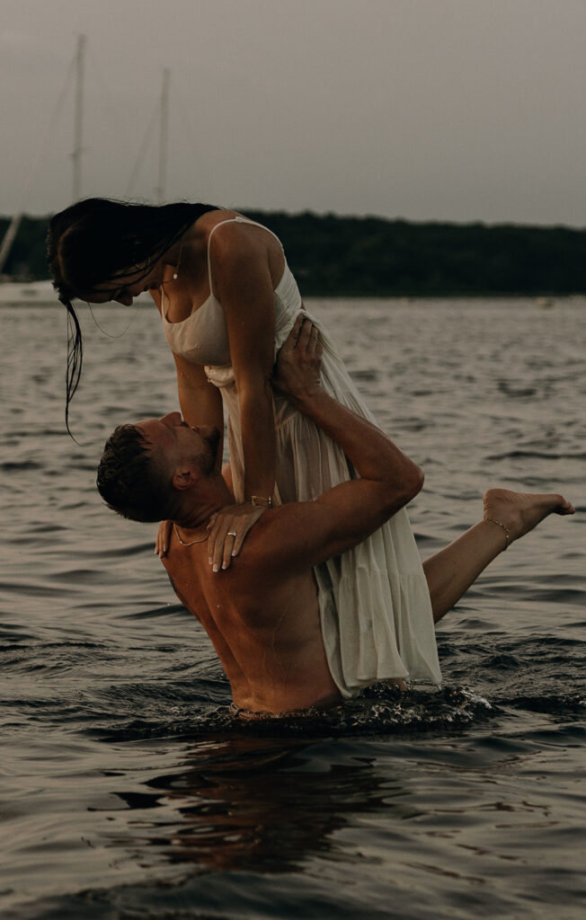 Adventure Wedding and Elopement Photographer New England | Aisha Lee Photography Cinematic Wedding Photographer | A couple stands in a lake at sunset. The man lifts the woman as they embrace joyfully. The tranquil water reflects their silhouette and the soft evening light.