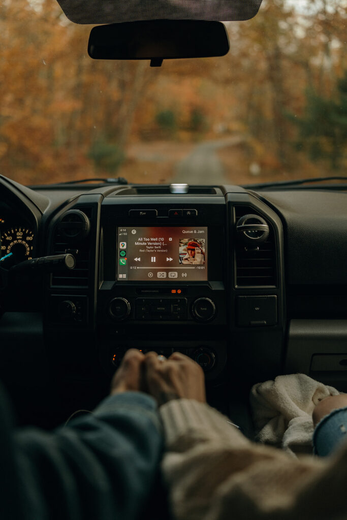 Destination Engagement Photographer New England | Aisha Lee Photography Cinematic Wedding Photographer | Inside a car, a couple holds hands near the console showing a music playlist, with a scenic autumn road visible through the windshield.