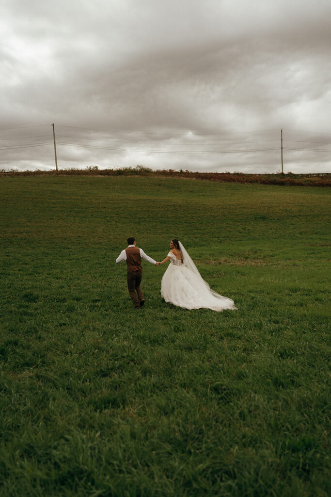Cinematic Wedding Photographer Rhode Island | Aisha Lee Photography Cinematic Wedding Photographer | A bride and groom walk hand-in-hand across a lush green field under a cloudy sky.