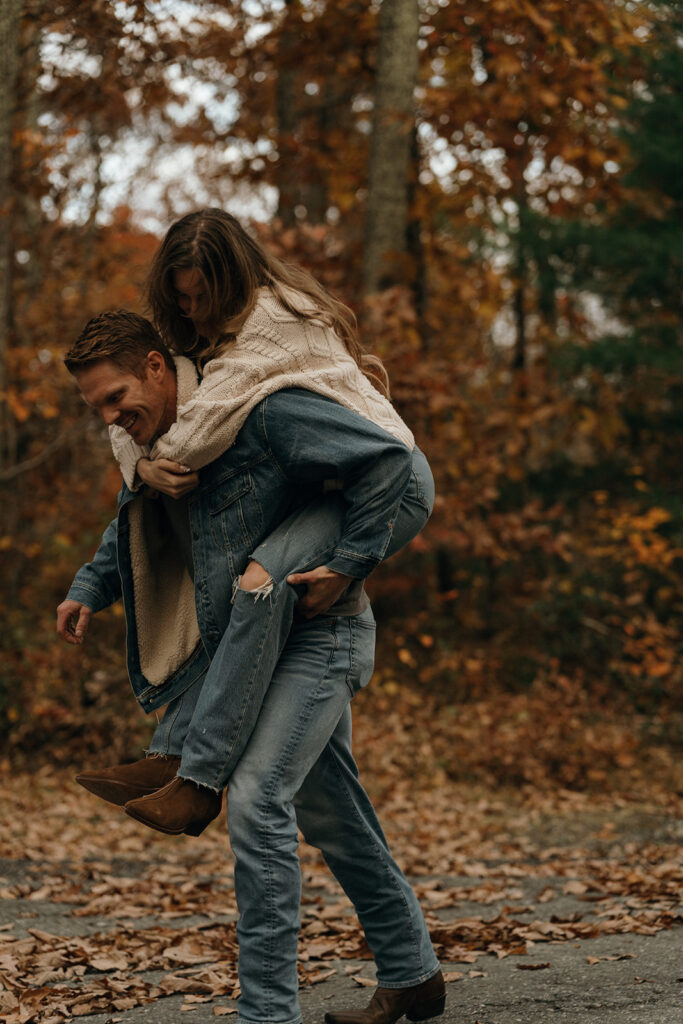 Adventure Wedding and Elopement Photographer New England | Aisha Lee Photography Cinematic Wedding Photographer | Man giving a woman a piggyback ride on a leaf-covered path, surrounded by autumn trees. Both wear denim and sweaters