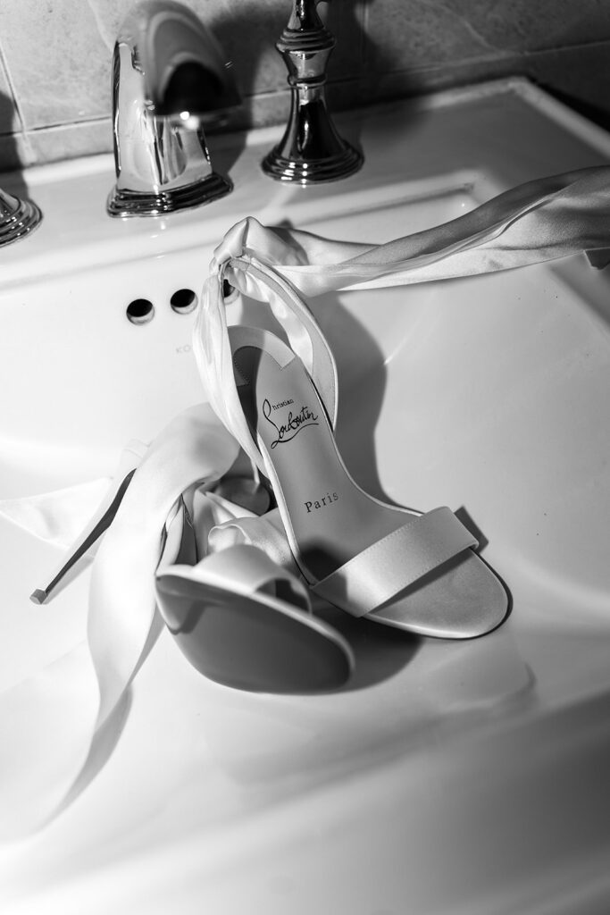 Black-and-white detail image of bridal heels resting on a sink beside soft ribbon accents and timeless accessories for a luxury New England wedding day.