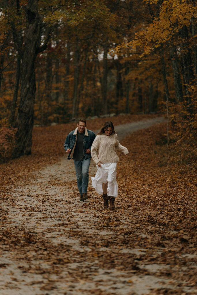 Cinematic Wedding Photographer Rhode Island | Aisha Lee Photography Cinematic Wedding Photographer | A couple walks joyfully along a leaf-covered path in an autumn forest.