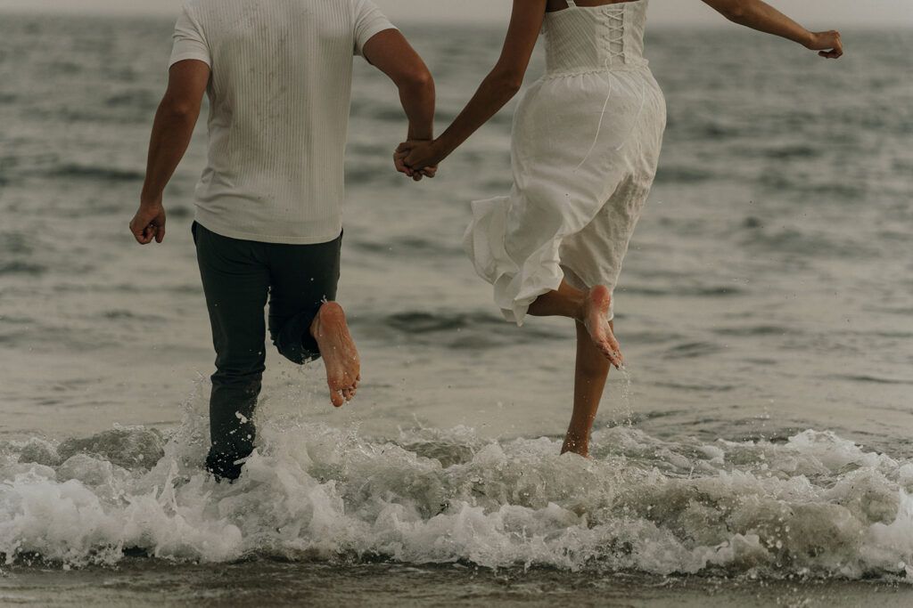 Oceanfront Weddings New England | Aisha Lee Photography Cinematic Wedding Photographer | A couple, holding hands, joyfully runs at the edge of the ocean. Their feet kick up water, conveying a carefree, romantic seaside moment.