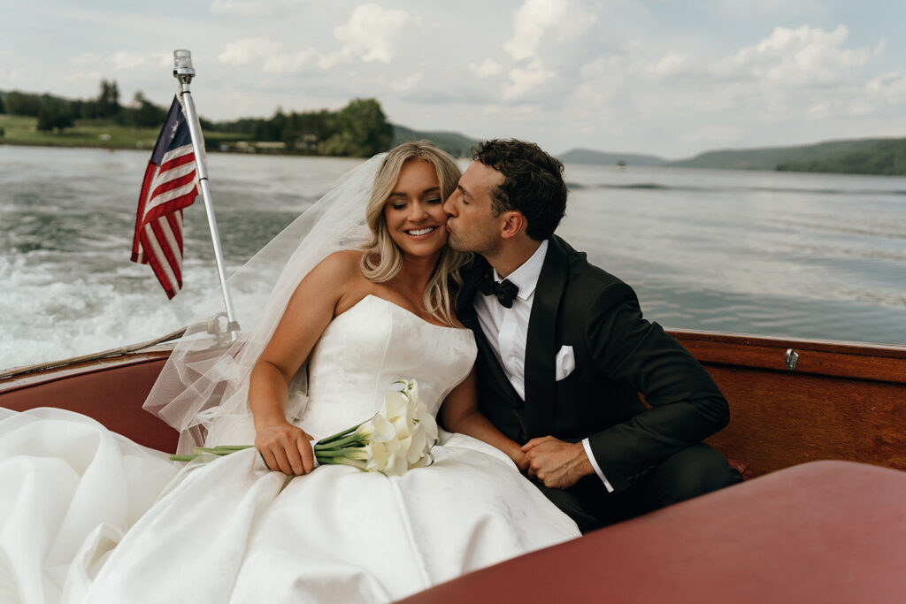 Rhode Island Wedding Photographer | Aisha Lee Photography Cinematic Wedding Photographer | A bride and groom are seated on a boat. The groom, in a black tuxedo, kisses the bride, who smiles and holds a bouquet of white flowers. The background shows a lake and trees under a cloudy sky, conveying a joyful and romantic atmosphere.