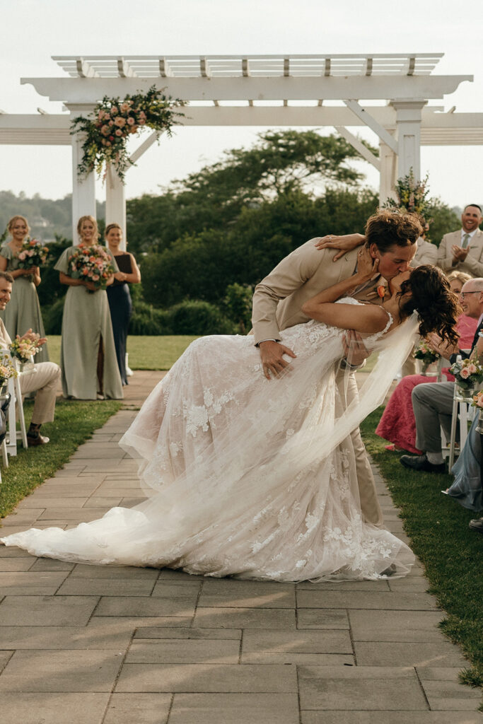 The groom dips the bride during an outdoor ceremony beneath a white pergola as guests look on, capturing a joyful and refined moment from a romantic New England castle wedding.