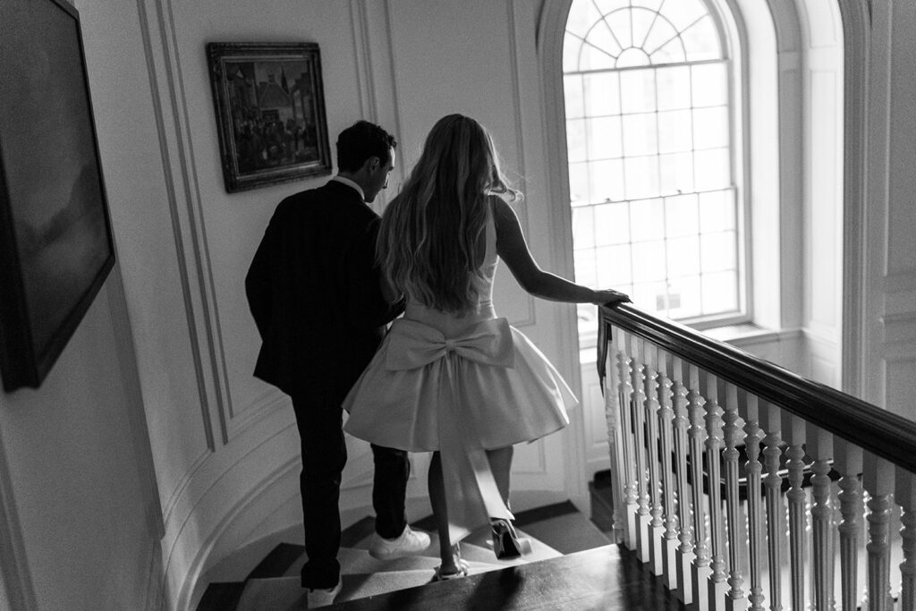 Unique Wedding Venues in New England | Aisha Lee Photography Cinematic Wedding Photographer | Black and white image of a couple descending stairs. The woman in a dress with a large bow, evokes elegance, while the man in a suit and sneakers adds a modern touch.