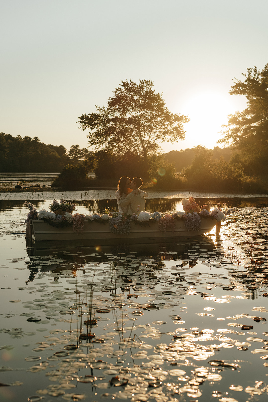 Destination Wedding Photographer Rhode Island | Aisha Lee Photography Cinematic Wedding Photographer | A couple embraces in a flower-adorned boat on a serene lake at sunset. They are surrounded by trees with a warm glow reflects on water