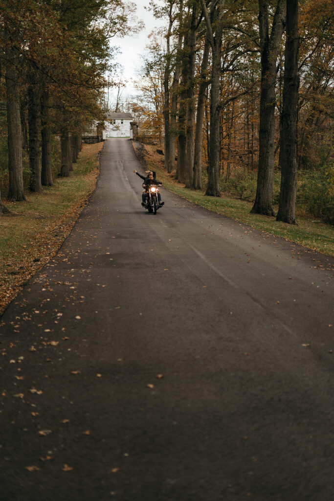 Unique Wedding Venues in New England | Aisha Lee Photography Cinematic Wedding Photographer | A motorcyclist rides down an empty tree-lined road, surrounded by autumn foliage. The scene feels adventurous and serene, leading towards a distant house.