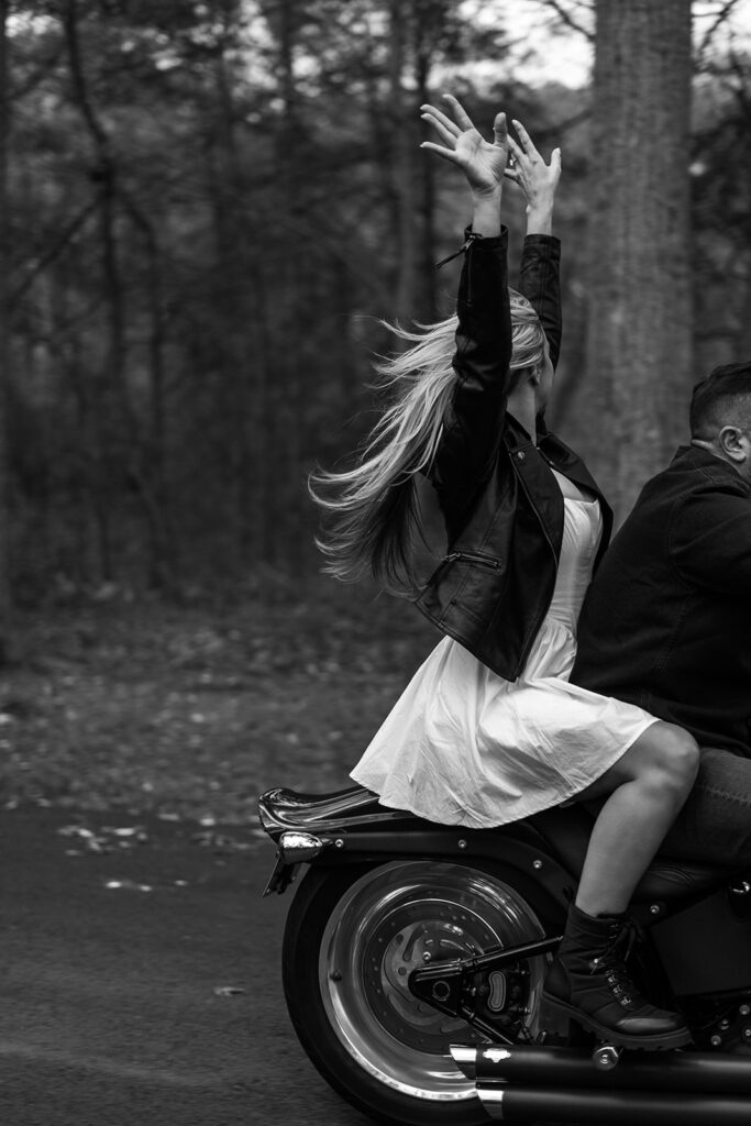 Bride throwing her hands in the air while riding on the back of a motorcycle through the woods during an adventurous celebration for couples choosing to elope in New England.