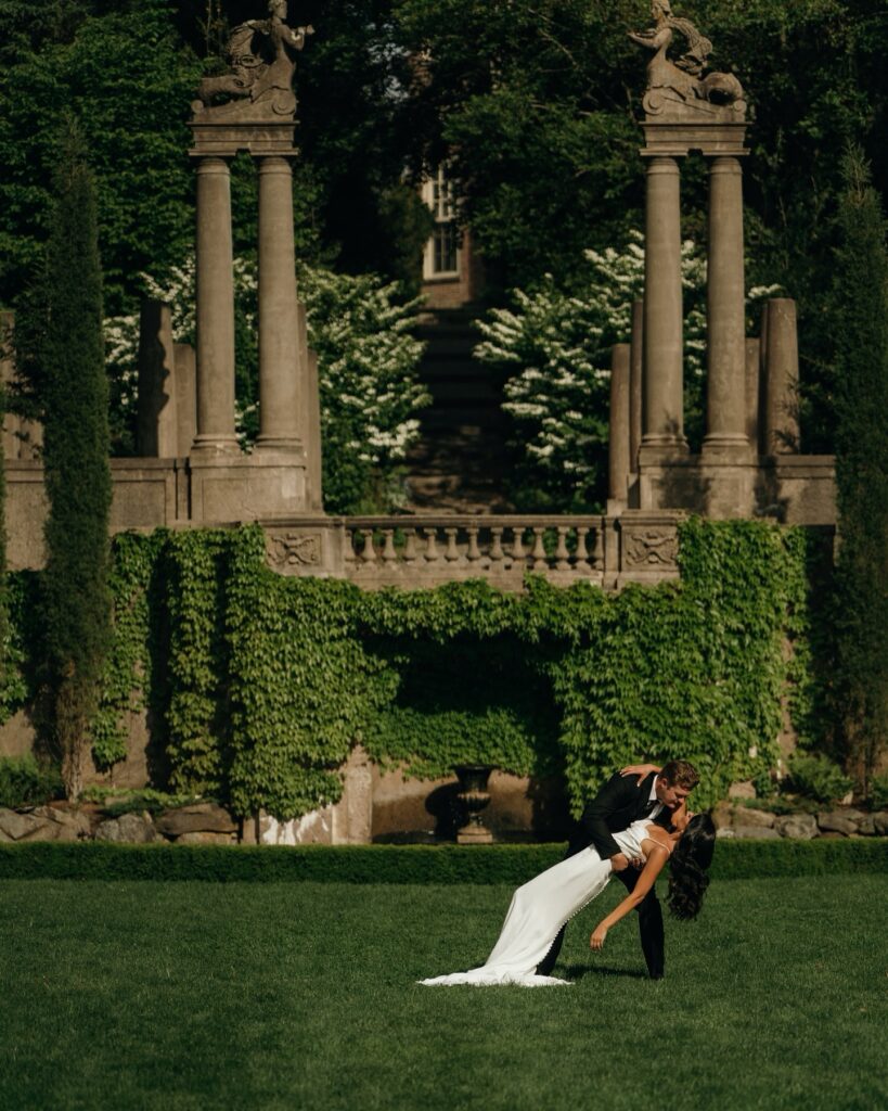 Castles in New England | Aisha Lee Photography Cinematic Wedding Photographer | A couple dances in front of ivy-covered stone columns in a lush garden. The man dips the woman, evoking romance and elegance on a sunny day.