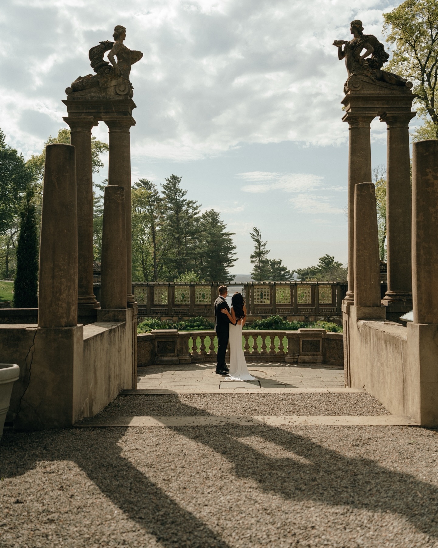 Castle Wedding Venues in New England | Aisha Lee Photography Cinematic Wedding Photographer | A couple in formal attire embraces under tall stone pillars in a serene garden setting. Soft light casts long shadows, evoking a romantic and timeless mood.