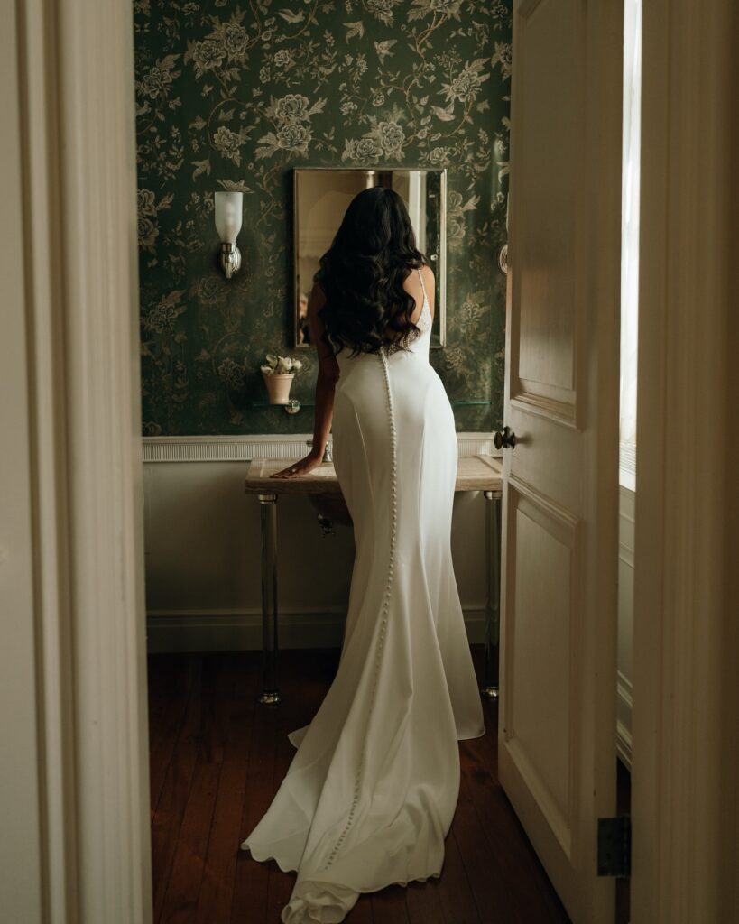 A bride stands at a vintage vanity adjusting her look, framed by patterned wallpaper and soft window light, reflecting the quiet elegance found in the best wedding venues in New England.