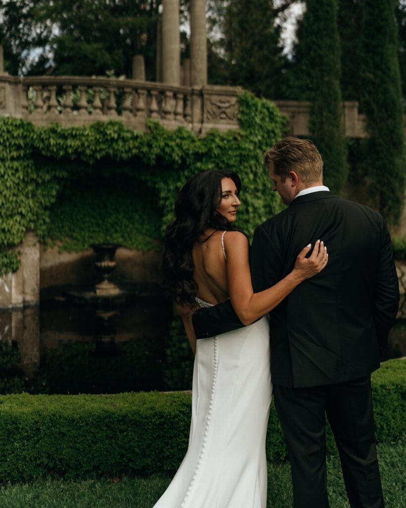 Rhode Island Wedding Photographer | Aisha Lee Photography Cinematic Wedding Photographer | A couple stands embrace in an elegant garden. The woman wears a white dress; the man is in a black suit. Ivy-covered walls and a serene pond are behind them.